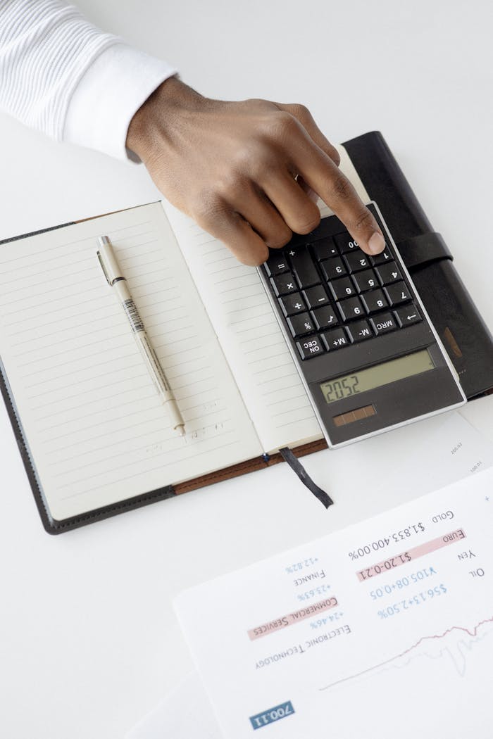 A hand using a calculator with financial documents and notebook, depicting an office setup for financial analysis.